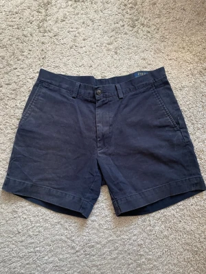Mörkblåa shorts från Polo Ralph Lauren - Klassiska mörkblå shorts från Polo Ralph Lauren med raka ben, sidofickor och bakfickor med knappar. Perfekta för sommaren och tillverkade i mjuk bomull. Snygg och tidlös design med bälteshällor och knappgylf. Kolla på alla bilder noga❗️