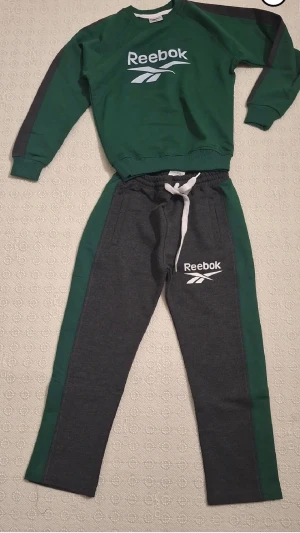 Grön och grå Reebok träningsdress - Snygg tvådelad träningsdress från Reebok i grönt och mörkgrått. Setet består av en sweatshirt med Reebok-logga på bröstet och matchande byxor med logga och dragsko i midjan. Perfekt för träning eller chill. Materialet känns mjukt och bekvämt.