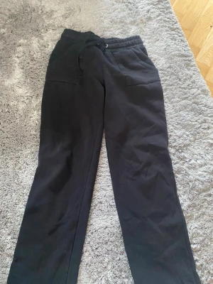 Svarta mjukisbyxor med dragsko - Säljer ett par svarta mjukisbyxor med raka ben och dragsko i midjan. Byxorna har två fickor framtill och är gjorda i ett mjukt och bekvämt material, perfekta för chill dagar. Passar dig som gillar en avslappnad stil.