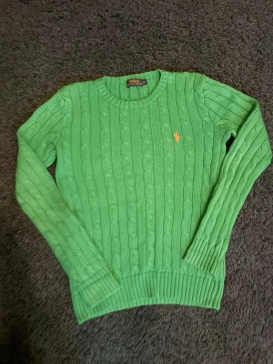 Ralph Lauren grön kabelstickad tröja - Snygg grön kabelstickad tröja från Polo Ralph Lauren med rund halsringning och det klassiska broderade logot på bröstet. Tröjan har långa ärmar och ribbade muddar vid ärmslut och nederkant. Mycket fint skick 