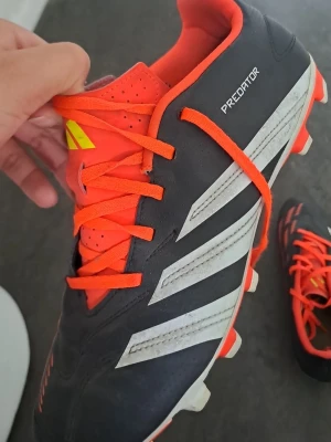 Adidas Predator fotbollsskor svart/orange - Säljer ett par Adidas Predator fotbollsskor i svart med vita ränder och orange detaljer. Skorna har snörning, låg profil och är tillverkade i syntetmaterial. Perfekta för dig som vill ha kontroll och stil på planen.