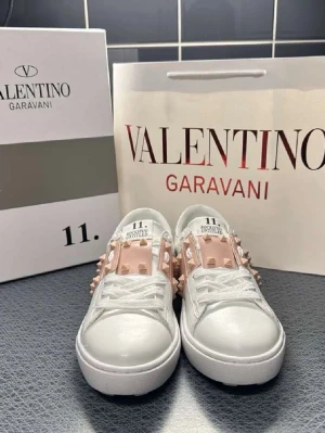 Valentino Garavani vita sneakers med nitar - Snygga vita sneakers från Valentino Garavani med ljusrosa band och ikoniska pyramidformade nitar. Skorna har en chunky sula och är tillverkade i slätt läder med klassisk snörning. Perfekta för dig som vill sticka ut med en lyxig och trendig look.