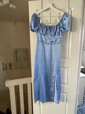 Ljusblå satinklänning - Söt ljusblå klänning med små vita blommor, puffärmar och slits framtill. Klänningen har en smickrande v-ringad ringning.