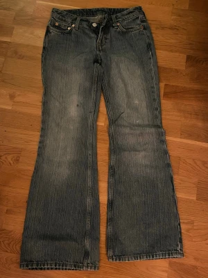 Blå bootcut jeans med slitningar - Säljer ett par blå bootcut jeans från Weekday med snygga slitningar och klassisk femficksdesign. Jeansen har en något utsvängd passform och detaljer i kopparfärgade nitar. Perfekta för en avslappnad och trendig look.