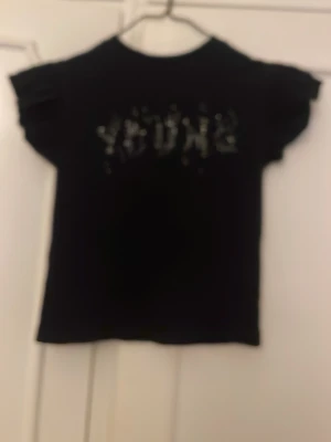 Svart t-shirt med pärlor - Cool svart t-shirt med korta ärmar och dekorativa pärlor som bildar texten 'YOUNG' på bröstet. Perfekt för dig som vill ha en enkel men ändå unik look. Materialet känns mjukt och bekvämt, och pärlorna ger en extra touch.