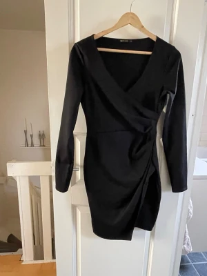 Svart omlottklänning med v-ringning - Stilren svart omlottklänning från Nelly med v-ringad hals och långa ärmar. Klänningen har en kort längd och markerad midja med draperad effekt på sidan. Perfekt för dig som gillar en elegant och enkel look.