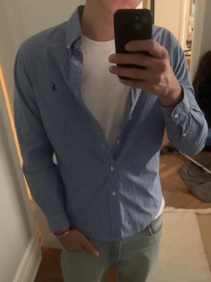 Blårutig skjorta från Ralph Lauren - Säljer en klassisk blårutig skjorta från Ralph Lauren med button-down krage och vita knappar. Skjortan har lång ärm och den ikoniska lilla loggan broderad på bröstet. Perfekt till jeans eller chinos för en clean och stilren look. Bröstmått: 47 cm Längd: 70 cm  