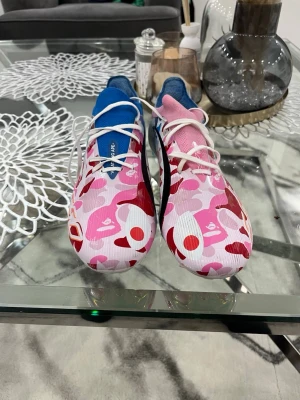 Adidas X BAPE rosa/blå fotbollsskor - Unika Adidas X BAPE fotbollsskor med färgstarkt camomönster i rosa, rött, vitt och blått. Ena skon har blå detaljer och den andra rosa, båda med vita skosnören och låg profil. Tillverkade i syntetmaterial för bästa grepp och känsla på planen.