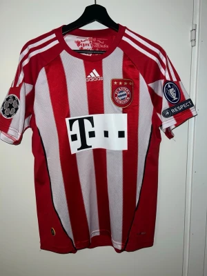 Bayern München matchtröja Adidas - Röd och vit randig Bayern München matchtröja från Adidas med Champions League-patchar på ärmarna och klubbmärke på bröstet. Tröjan har Telekom-logga på magen och klassiska Adidas-ränder på axlarna. Tillverkad i ventilerande funktionsmaterial.