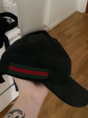 Svart keps från Gucci med röd/grön detalj - Säljer en svart keps från Gucci med den klassiska röd-gröna randen på sidan. Kepsen har böjd skärm och justerbar passform. Tillverkad i ett slitstarkt tyg som känns lyxigt och stilrent. Perfekt för dig som vill ha en exklusiv accessoar med ikonisk design.