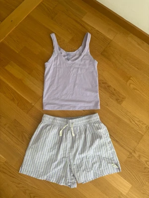 Ljuslila pyjamas-set linne & shorts - Säljer ett mjukt och bekvämt pyjamas-set från H&M. Linnet är ljuslila med breda axelband och rund halsringning. Shortsen är vita med tunna blå ränder och har resårmidja samt dragsko för skön passform. Perfekt för chill dagar hemma.