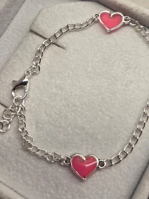 Armband med rosa hjärtan som lyser - Snyggt silverfärgat armband i rostfritt stål med två rosa hjärtan i emalj som lyser lila i mörker. Kedjan är tunn och har ett klassiskt karbinlås. Perfekt accessoar för dig som gillar unika detaljer och färgklickar i din stil. Att vara ute i dagsljus en kort stund räcker för att hjärtanen ska börja lysa i mörkare omgivning. Har skapat detta unika armband för hand inklusive färg och glow pulver. Tål vatten och missfärgas inte. Lång livslängd. 