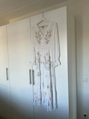 Vit långklänning med blommor - Superfin vit långklänning med broderade blommor i flera färger. Klänningen har v-ringning, lång ärm med lätt volang vid ärmslut, knappar framtill och en slits. Perfekt för sommarens alla tillfällen och ger en romantisk vibe. Endast provat klänningen. 