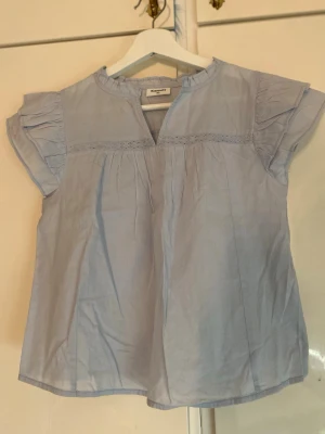 Ljusblå blus från Kappahl  - Ljusblå blus från Kappahl med korta ärmar och volangdetaljer. Blusen har en rund halsringning med liten v-ringning framtill och dekorativ spets längs bröstet. Materialet är tunt och lätt, perfekt för varma dagar.🩵☀️