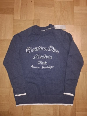 Stickad tröja D10R - Exklusiv blå stickad tröja från Christian D10R med vit broderad text på bröstet. Tröjan har rund hals, ribbade muddar och vita detaljer vid ärmslut och nederkant. Perfekt för dig som vill ha en lyxig och snygg look med Paris-vibe.                             Storlek är S men skulle säga att det pssar mer som M!