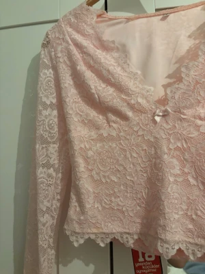 Ljusrosa spetsblus med v-ringning - Säljer en ljusrosa blus i spets med v-ringning och långärmade ärmar. Blusen har en liten rosett framtill och fina spetsdetaljer längs nederkanten. Perfekt för dig som gillar romantiska och feminina toppar.