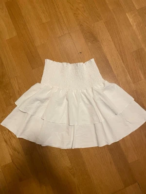 Vit volangkjol med smock - Köpt i Spanien, Säljer en söt vit kortkjol med bred smock i midjan och dubbla volanger. Kjolen har en luftig och flowy känsla, perfekt för varma dagar. Passar till många olika stilar och är superenkel att matcha med olika toppar.