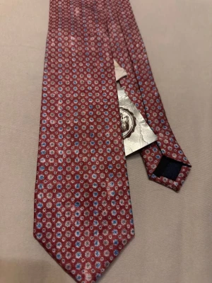 Röd slips med småblommigt mönster - Snygg röd slips från Portico med småblommigt mönster i vitt och blått. Tillverkad i ett glansigt tyg som ger en exklusiv känsla. Perfekt accessoar för att lyfta din outfit med lite extra färg och stil.