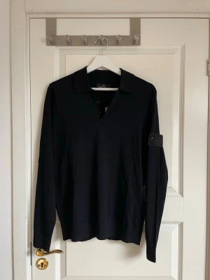 Svart långärmad topp Stone Island - Svart långärmad topp från Stone Island med klassisk krage och knappdetalj vid halsen. Tillverkad i mjuk bomull med lite elastan för extra komfort. Ikonisk logopatch på ärmen ger en stilren touch. Perfekt för dig som gillar minimalistisk och clean stil.
