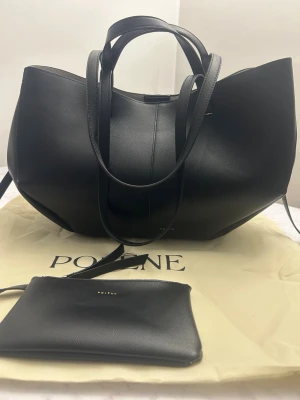 Svart handväska från Polène - Stilren svart handväska från Polène med elegant och modern form. Väskan har både korta och långa handtag, vilket gör den enkel att bära på olika sätt. Tillverkad i slätt skinn med minimalistisk design och diskret logga framtill. Den är 32 x 38 x 17 cm. 