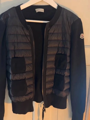 Svart cardiganjacka från Moncler - Snygg svart cardiganjacka från Moncler med quiltad front och mjuka stickade ärmar. Jackan har dragkedja framtill, två fickor och Moncler-logga på ärmen. Perfekt för lager på lager och en clean look.