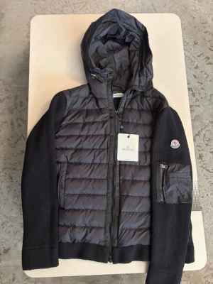 Moncler cardigan  - Snygg svart pufferjacka från Moncler med stickade ärmar och huva. Jackan har quiltad front, dragkedja, fickor och en patch med Moncler-logga på ärmen. Perfekt för kalla dagar och riktigt trendig look. Pris kan diskuteras. 