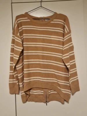 Beige randig stickad tröja med dragkedja - Stickad tröja i beige med vita horisontella ränder. Tröjan har rund halsringning, långa ärmar och en unik dragkedja längs ryggen. Avslappnad passform och ribbade muddar vid ärmslut och nederkant. Perfekt för en chill och trendig look.