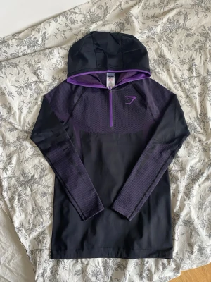 Gymshark Onyx V5 Lila hoodie - Snygg svart hoodie från Gymshark med lila detaljer och halv dragkedja framtill. Tröjan har huva, långa ärmar och ett mönstrat tyg över bröstet och armarna. Perfekt för träning eller chill. Tillverkad i ett stretchigt funktionsmaterial.