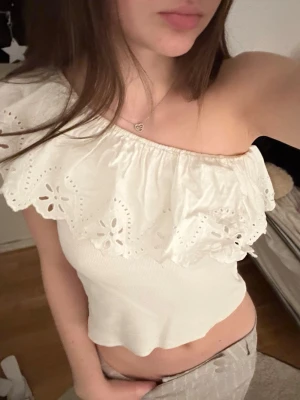 Vit offshoulder topp med volang - Söt vit offshoulder topp med bred volangkant och broderade spetsdetaljer. Toppen är ärmlös och har en figurnära passform med lätt ribbad struktur. Perfekt för dig som vill ha en romantisk och trendig look.