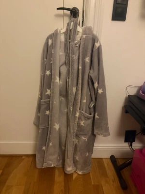 Grå morgonrock med stjärnor och huva - Mysig grå morgonrock med vita stjärnor, huva och två stora fickor framtill. Tillverkad i mjuk fleece och har ett matchande midjebälte. Perfekt för att chilla hemma och hålla sig varm under kalla morgnar.