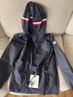 Moncler Jacka - Helt ny snygg moncler vindjacka  med huva och dragkedja framtill. Jackan har vita och röda ränder på huvan samt Moncler-logga på ärmen. Tillverkad i lätt, vattenavvisande material och har praktiska fickor med tryckknapp. Nfc- tag finns prislapp kvar. Storlek M men passar S med. Säljs för 1400kr!