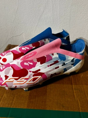Adidas x BAPE F50 fotbollsskor - Unika Adidas x BAPE F50 fotbollsskor med färgstarkt kamouflagemönster i rosa, rött, blått och vitt. Skorna har en strömlinjeformad design utan snörning, med blå och rosa detaljer på ovandelen. Tillverkade i syntetmaterial för bästa prestanda på planen.