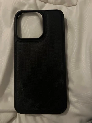 Svart mobilskal i syntetmaterial - Snyggt svart mobilskal till iPhone 13 Pro med stilren design och texturerad yta. Skalet har en stor öppning för kameran och är tillverkat i tåligt syntetmaterial som skyddar mobilen mot repor och stötar. Passar perfekt för dig som vill ha ett enkelt och diskret skydd till din telefon.