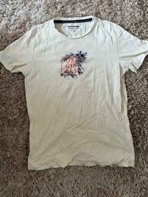 Vit t-shirt Jack & Jones Originals - Vit t-shirt från Jack & Jones Originals med tryck framtill i form av blå blad och orange texten 'ORIGINALS' i en fyrkant. Klassisk rund hals och korta ärmar. Tillverkad i mjuk bomull, perfekt för en avslappnad stil.