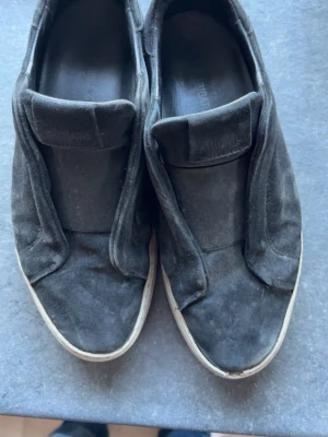 Arigatos - Svarta slip-on sneakers med ovandel i mocka och vit sula. Skorna har en enkel och stilren design utan snörning, vilket gör dem lätta att ta på och av. Passar perfekt till en avslappnad streetwear-look.