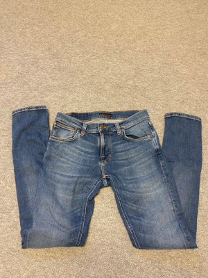 Blå jeans från Nudie Jeans - Snygga blå jeans från Nudie Jeans med klassisk femficksdesign och orangea sömmar. Jeansen har raka ben och en cool tvättad look med slitningar framtill. Bakfickorna har Nudie Jeans signaturbrodyr och läderpatch i midjan. Perfekta för en avslappnad stil.