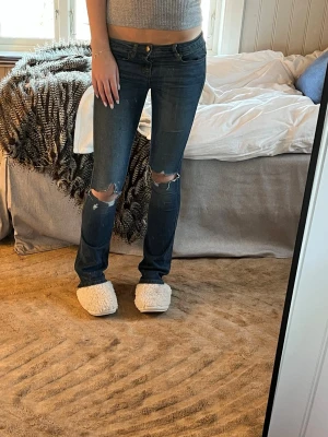 Mörkblå slitna jeans med hål - Säljer ett par mörkblå flaird jeans med slitningar vid knäna för en avslappnad och trendig look. Jeansen har klassisk femficksdesign och knappgylf. Perfekta för dig som gillar en casual och edgy stil.