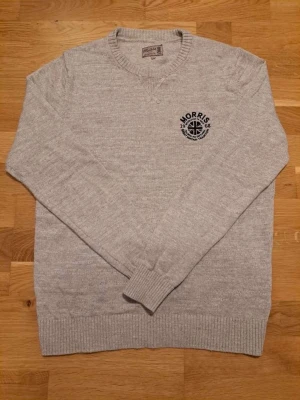  Morris tröja - Snygg ljusgrå stickad tröja från Morris med rund halsringning och svart broderad logga på bröstet. Tröjan har ribbade muddar vid ärmslut och nederkant.  Skriv vid några frågor!