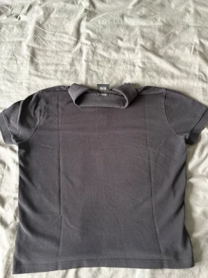 Mörkgrå pikétröja från BOSS - Säljer en stilren mörkgrå pikétröja från BOSS med klassisk krage och knappar framtill. Tröjan har korta ärmar och diskret BOSS-logga broderad på bröstet. Perfekt till jeans eller shorts för en clean look.