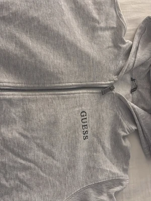 Grå hoodie med dragkedja från Guess - Snygg grå hoodie från Guess med dragkedja framtill och klassisk huva. På bröstet finns Guess-loggan broderad i svart och på ryggen ett stort rött och svart tryck med den ikoniska triangeln. Tillverkad i mjukt bomullsmaterial för skön känsla.