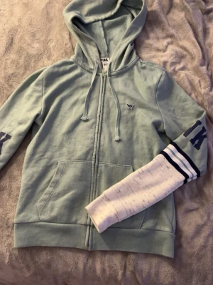 Mintgrön hoodie från PINK - Mintgrön hoodie med dragkedja från PINK Victoria's Secret. Har huva med dragsko, fickor framtill och ett litet hundmärke på bröstet. Ärmarna har mörkblå text och vita detaljer med ränder. Perfekt för en chill och sportig look.