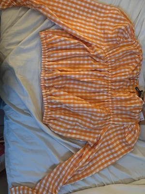 Orange rutig croppad blus - Superfin croppad blus i orange och vitt rutigt mönster. Blusen har långa ärmar med resår i midjan och ärmarna för en snygg puffig effekt. Perfekt till jeans eller kjol, så snygg orange/coral färg