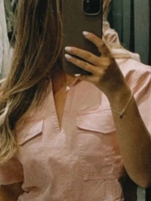 Ljusrosa croppad Jeans/Denim topp från Zara - Säljer en ljusrosa croppad topp från Zara med korta ärmar och två stora bröstfickor. Toppen har en v-ringad öppning och är gjord i ett mjukt bomullsmaterial. Perfekt för dig som gillar en chill och trendig stil. 100% Bomull.