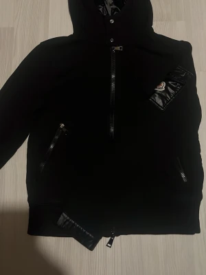 Svart hoodie från Moncler med huva - Svart hoodie från Moncler med dragkedjor och huva. Har en snygg detalj i form av en glansig ficka med Moncler-logga på ärmen. Köpt från room 1006, Perfekt för en stilren och sportig look.