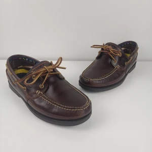 Timberland Earthkeepers Kiawah Bay Handsydda seglarskor, chokladbrunt, EU43 - Denna klassiska Timberland-seglarsko är tillverkad i ett slitstarkt, mörkbrunt fullnarvsläder med en traditionell handsydd konstruktion som ger både stil och hållbarhet. Skon kännetecknas av sitt funktionella 360-graders snörsystem med råhudssnören för en optimal passform samt en robust, räfflad gummisula som ger ett säkert grepp på våta underlag.