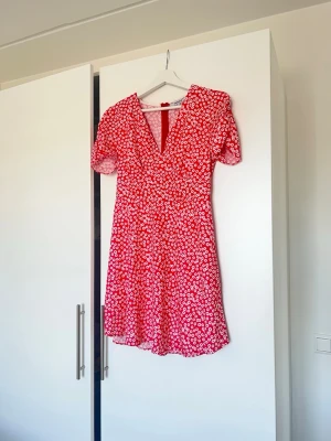 Röd blommig kortklänning Chiquelle - Söt röd kortklänning från Chiquelle med vitt blommönster, v-ringning och korta puffärmar. Klänningen har en figurnära passform upptill och lätt utställd kjol. Perfekt för sommarens alla tillfällen!