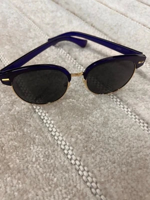 Blå clubmaster solglasögon - Snygga solglasögon i clubmaster-modell med mörka runda linser. Bågarna är djupblå med guldfärgade detaljer och metallram runt linserna. Klassisk och trendig design som passar till många stilar. Plast och metall i kombination för en cool look.