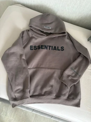 Brun Essentials Fear of God hoodie - Säljer en brun hoodie från Essentials Fear of God med svart tryck på bröstet och ärmen. Hoodien har en stor huva, ribbade muddar och en klassisk känguruficka framtill. Perfekt för dig som gillar streetwear och vill ha en clean look. Den är helt ny, från USA. För män och kvinnor. 
