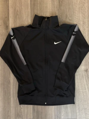 🖤Svart träningsjacka från Nike(Barn)🖤 - Snygg svart träningsjacka från Nike med hel dragkedja och hög krage. Jackan har vita Nike-loggor på bröstet och ärmarna samt vita prickiga detaljer längs axlar och armar. Tillverkad i ett mjukt och stretchigt material som passar perfekt till träning eller sport. Passar dig som är ca 150-160 cm. 