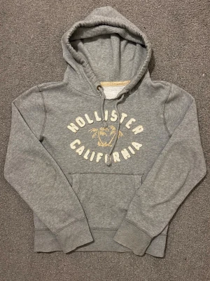Vintage Hollister Hoodie - Sällynt och Vintage Hollister Hoodie! | Palmträd i mitten av textryck centrerat på hoodien🌴| Mycket bra skick förutom en fläck på högra ärmen, syne ej mkt alls, se sista bild | Storlek S | Perfekt till hösten | Längd ≈60 cm, Bredd 53cm | Modell 175cm, 68kg Priset ej hugget i sten!